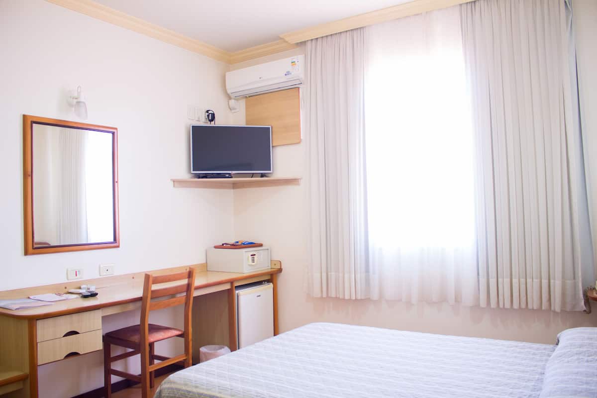 Una habitación de hotel con cama, escritorio, silla, televisor y aire acondicionado.