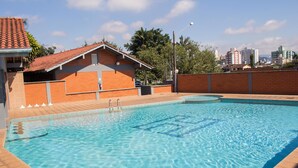Una piscina con bordeado de baldosas, un edificio de ladrillo con techo rojo y un paisaje urbano al fondo.