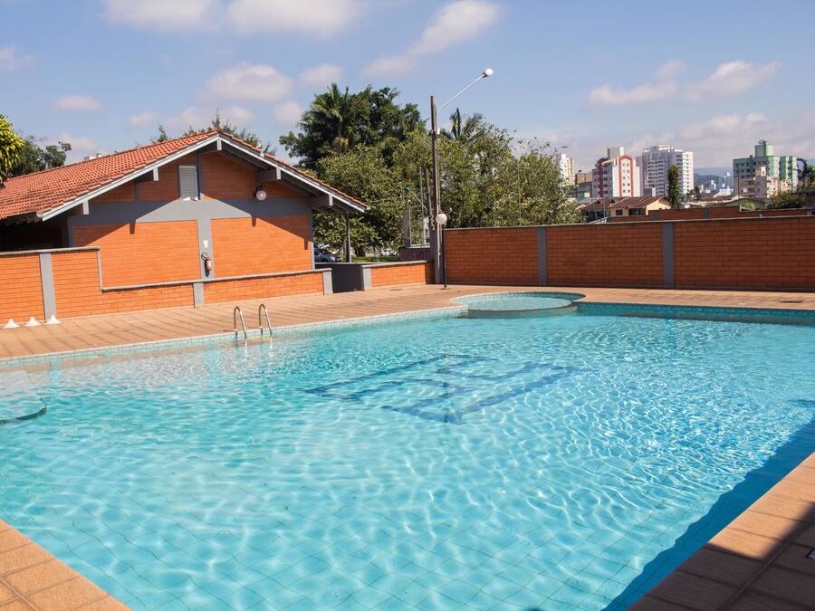 Una piscina con bordeado de baldosas, un edificio de ladrillo con techo rojo y un paisaje urbano al fondo.