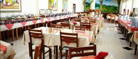 Uma sala de jantar com várias mesas e cadeiras, um balcão longo com alimentos e um mural na parede.