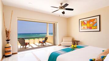 A bedroom with a large bed, a ceiling fan, a view of the ocean, and a framed abstract painting.