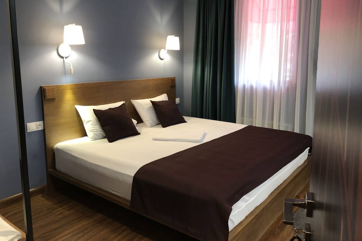 Ein ordentlich bezogenes Bett mit weißer und brauner Bettwäsche, zwei Wandlampen und ein Fenster mit durchsichtigen Vorhängen.