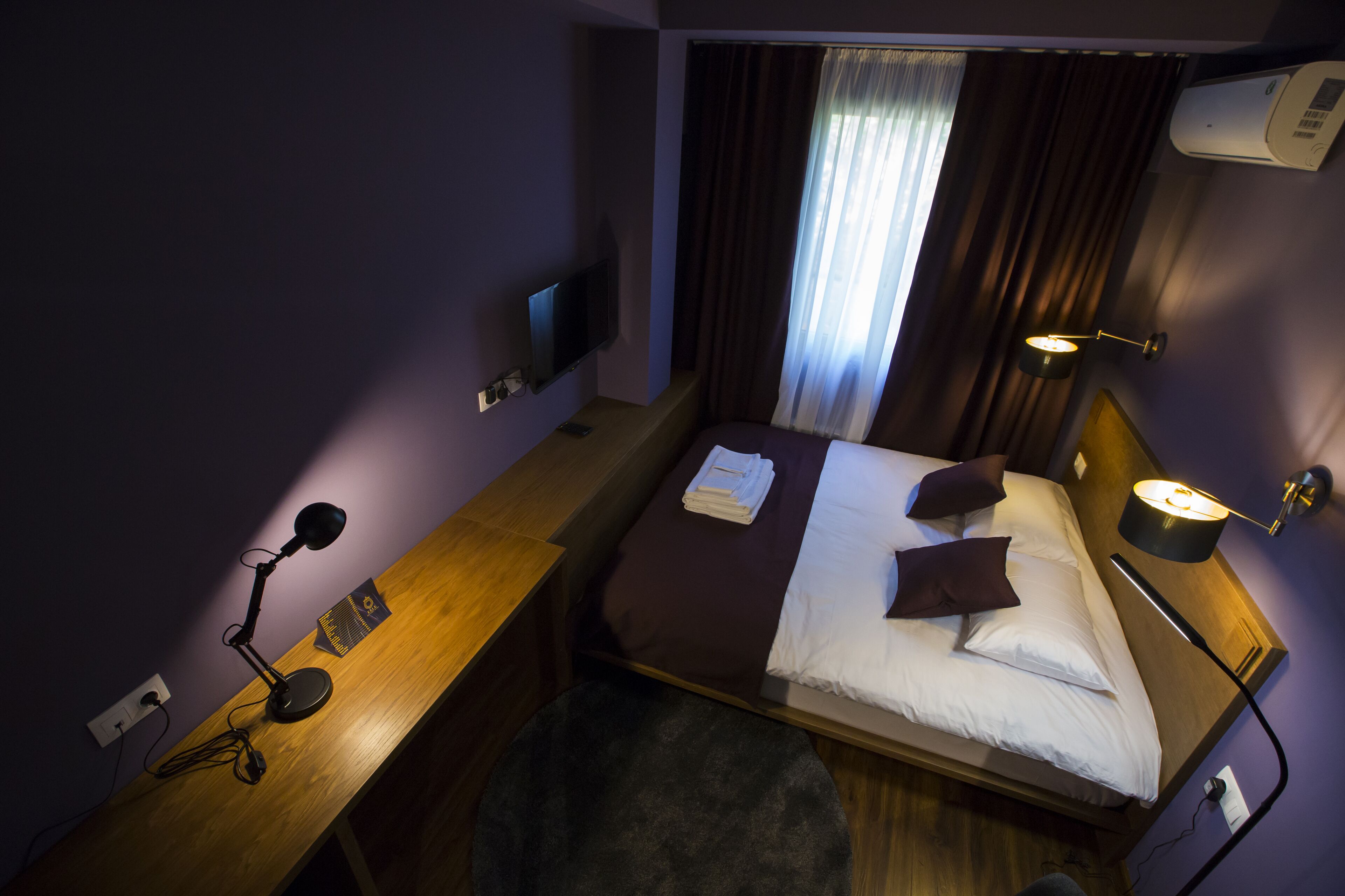 A hotel room with a bed, bedside lamps, a desk with a lamp, a TV, and a window with curtains.
