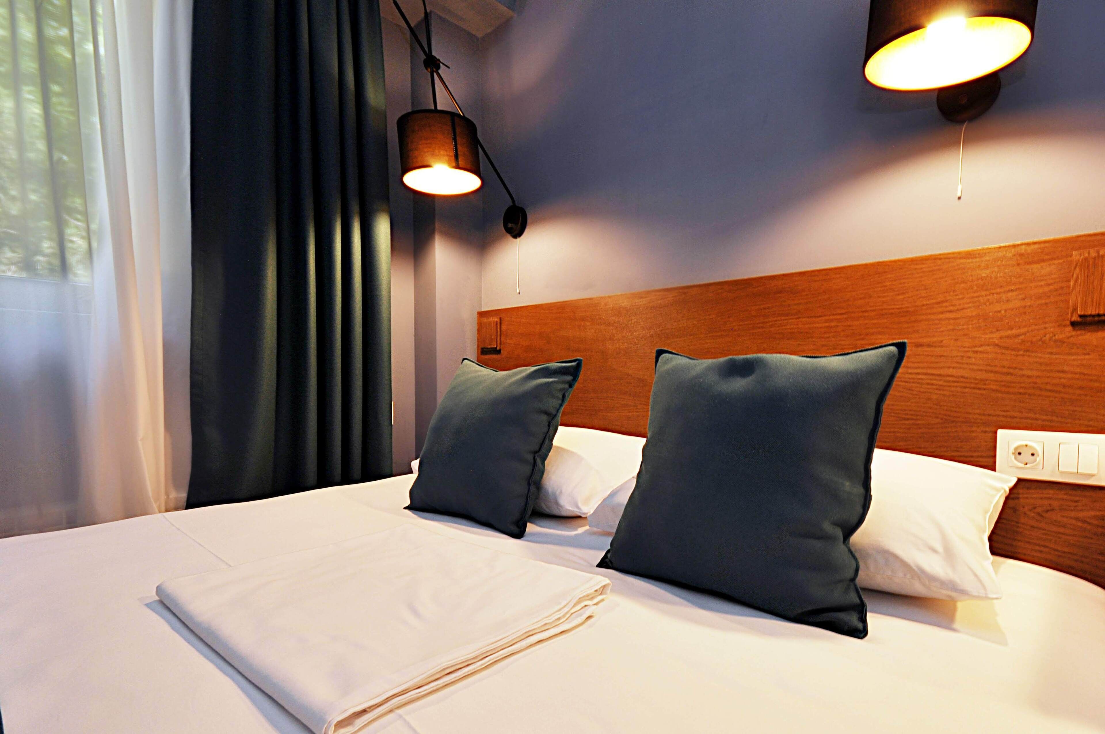 A neatly made bed with white linens and two dark pillows, a wooden headboard, and two wall-mounted lights.