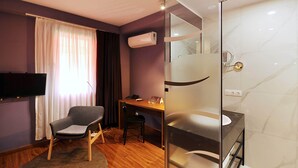 Une chambre d’hôtel moderne dotée d’une télévision à écran plat, d’un bureau, d’une chaise, d’une douche en verre et d’une salle de bain carrelée en marbre.