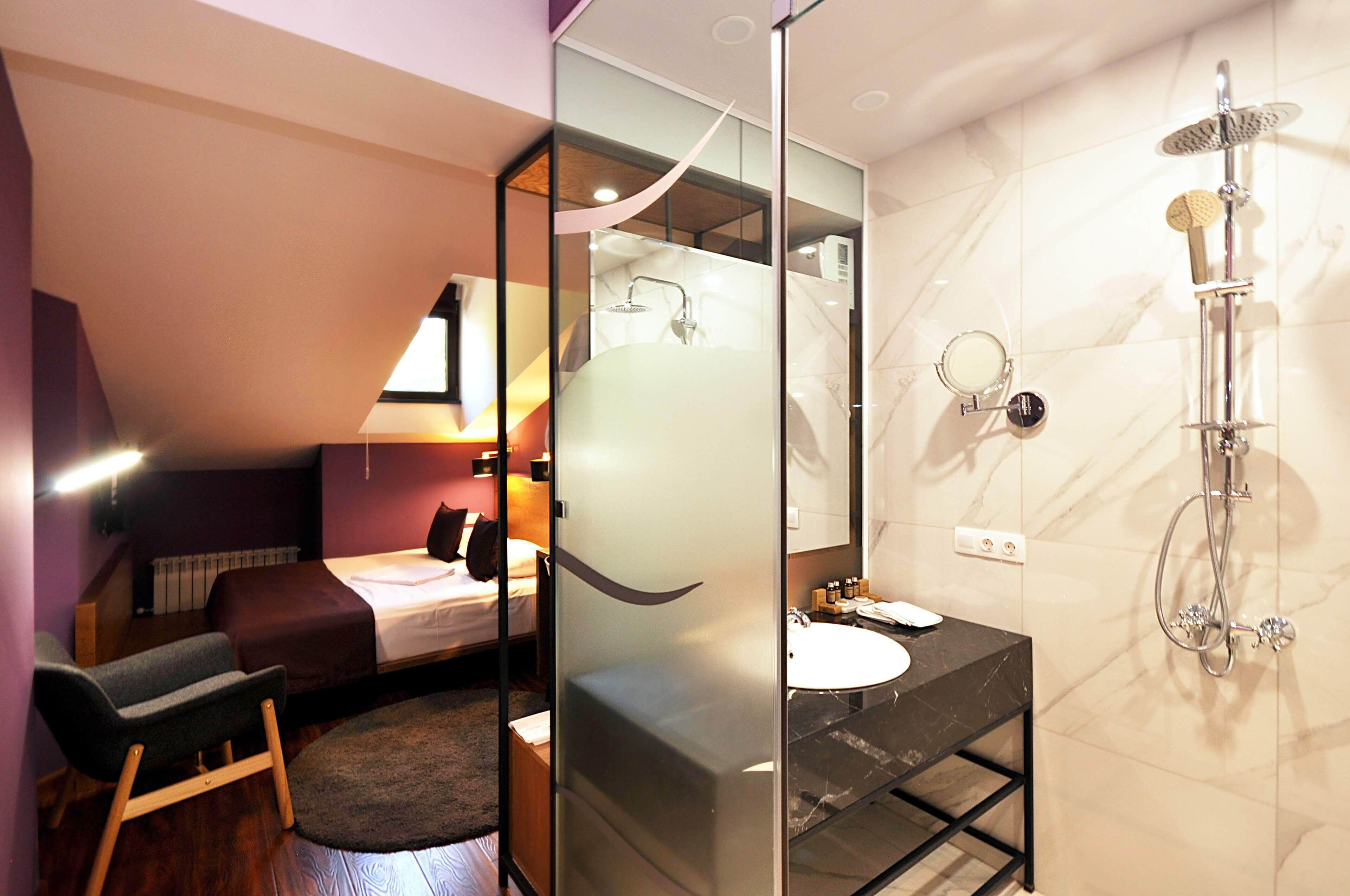 A modern hotel room with a glass partition separating the bedroom from the bathroom. The bedroom features a bed, a small seating area, and a desk. The bathroom includes a shower, a sink, and a mirror.