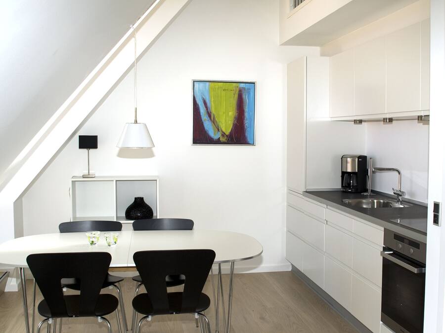 A modern kitchen with white cabinets, a dining table with two chairs, a coffee maker, and a painting on the wall.