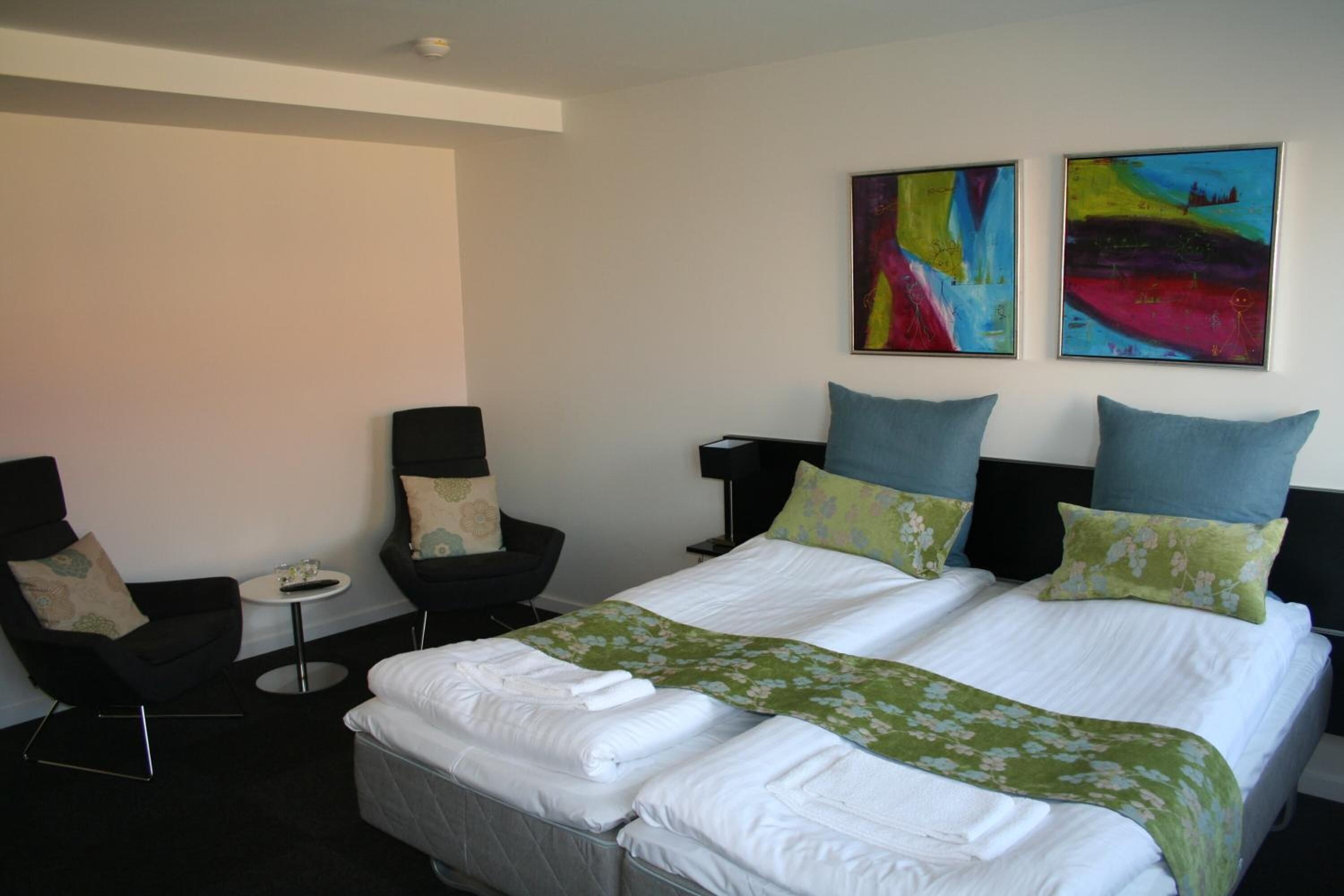 A hotel room with a large bed, two chairs, a small table, and two abstract paintings on the wall.