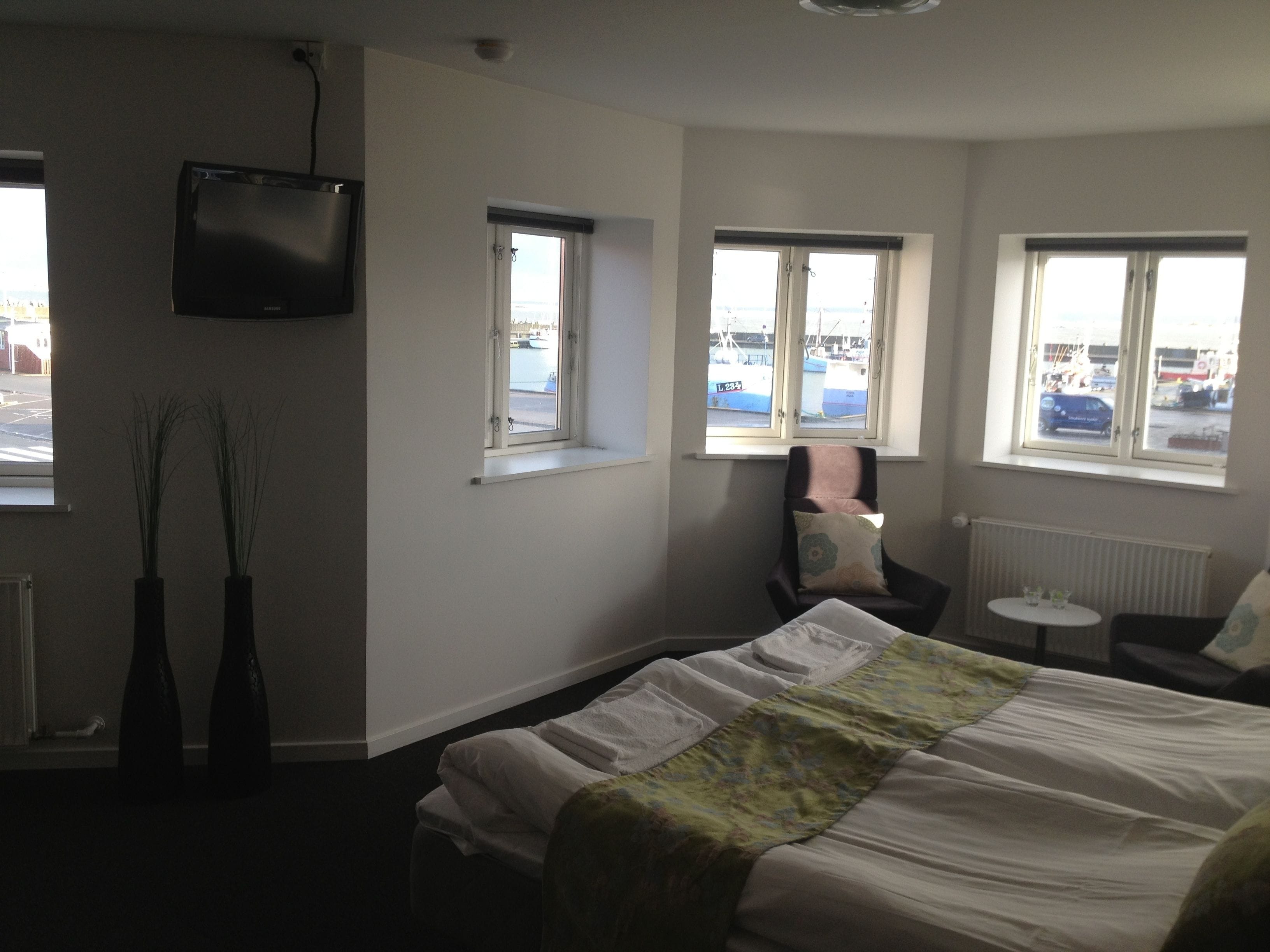 A hotel room with a bed, a chair, a small table, and two windows.