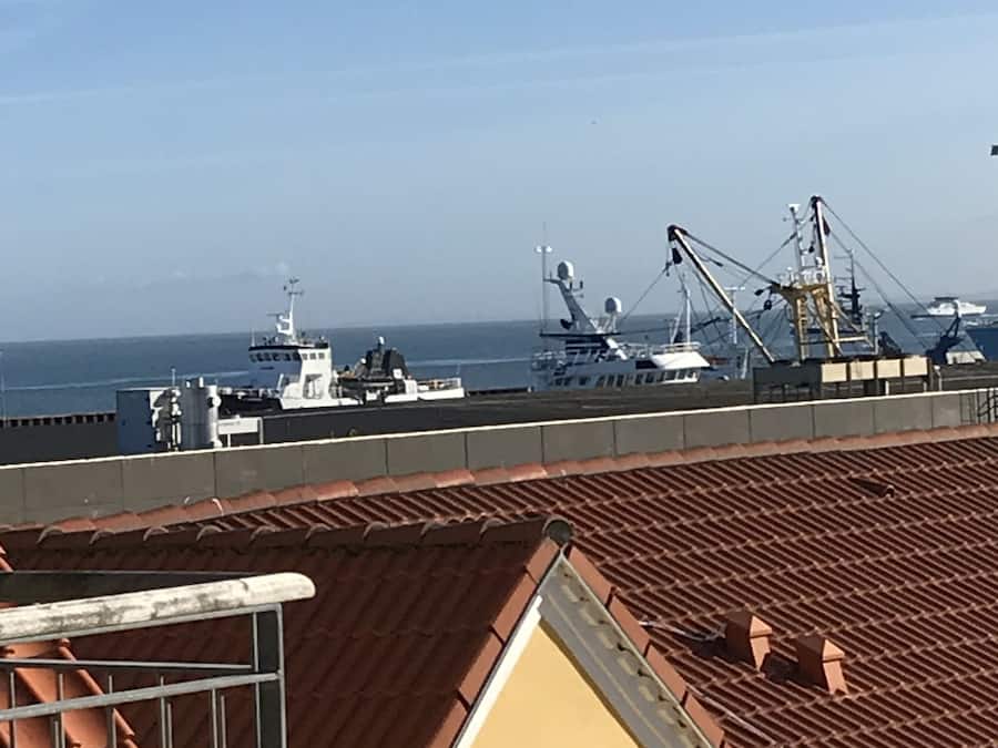 A view of a port with ships and cranes, a clear sky, and a building with a tiled roof in the foreground.