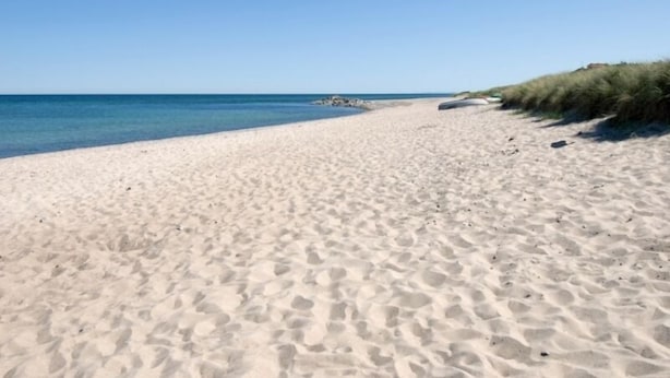 Een zandstrand met helderblauw water en een grasduin op de achtergrond.