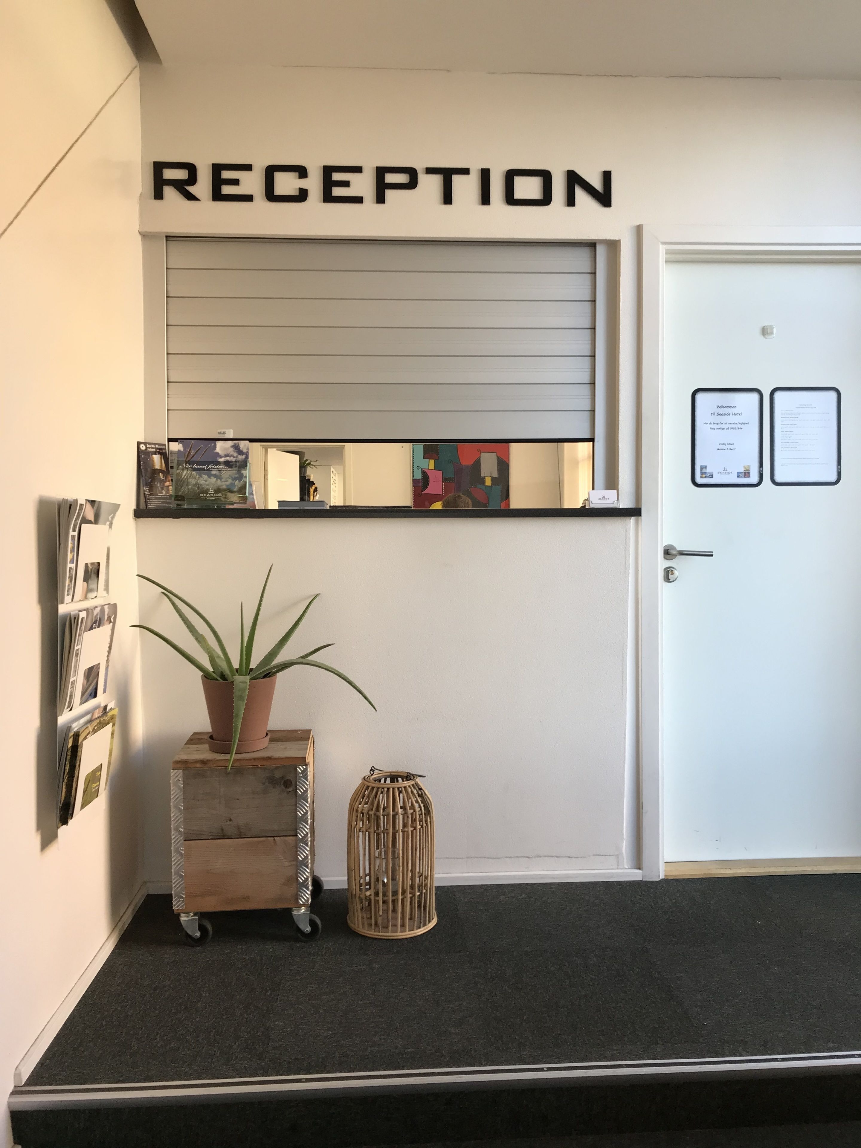 A reception area with a sign that reads "RECEPTION" above a door. There is a potted plant on a wooden cart and a decorative wicker basket on the floor.
