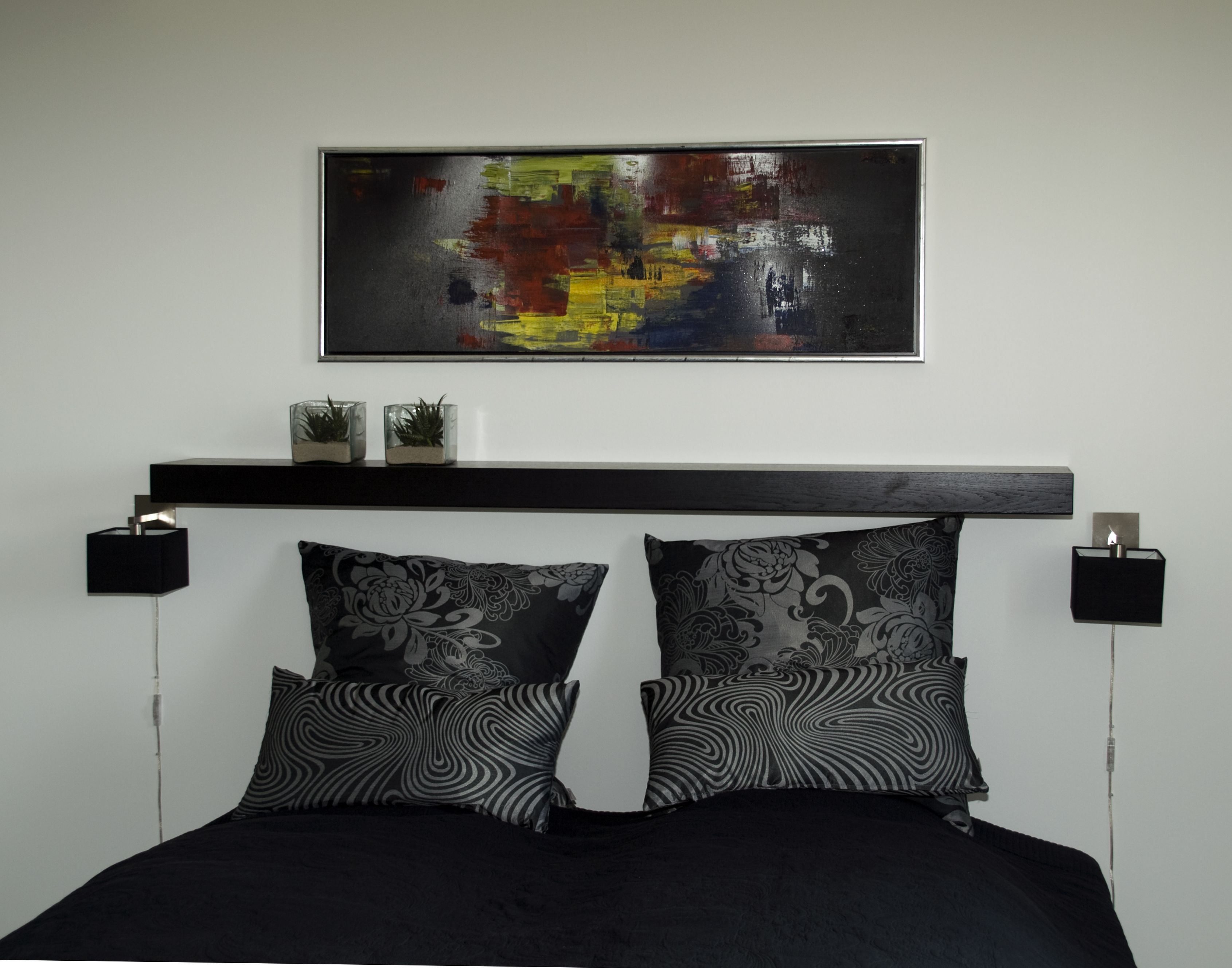 A modern bedroom with a black bed, two patterned pillows, a black headboard, a wall-mounted abstract painting, and two bedside lamps.