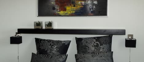 Un dormitorio moderno con una cama negra, dos almohadas estampadas, un cabecero negro, un cuadro abstracto colgado en la pared y dos lámparas de noche.