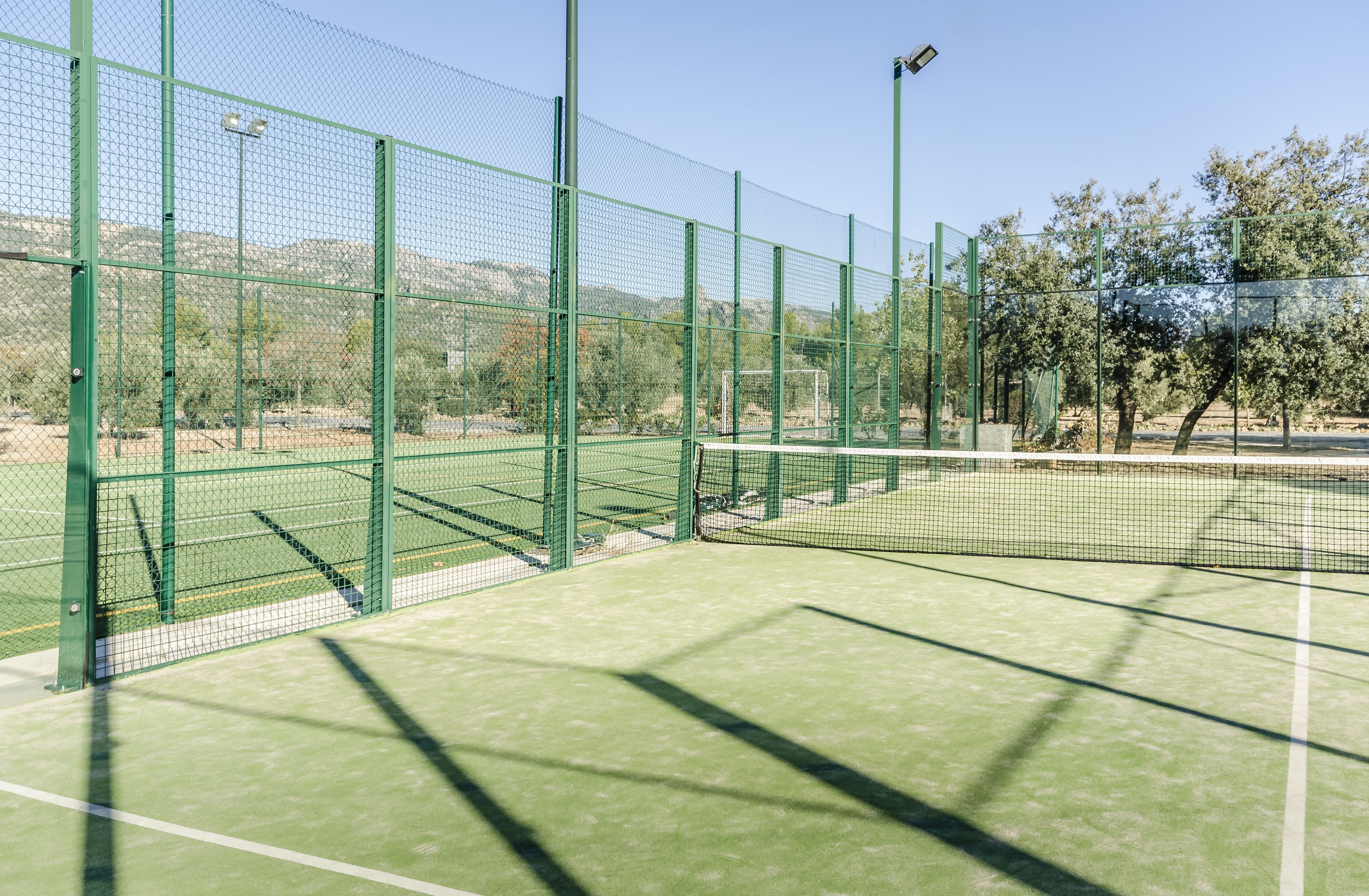 Pista de tenis con vallas verdes y una red, rodeada de árboles y colinas.