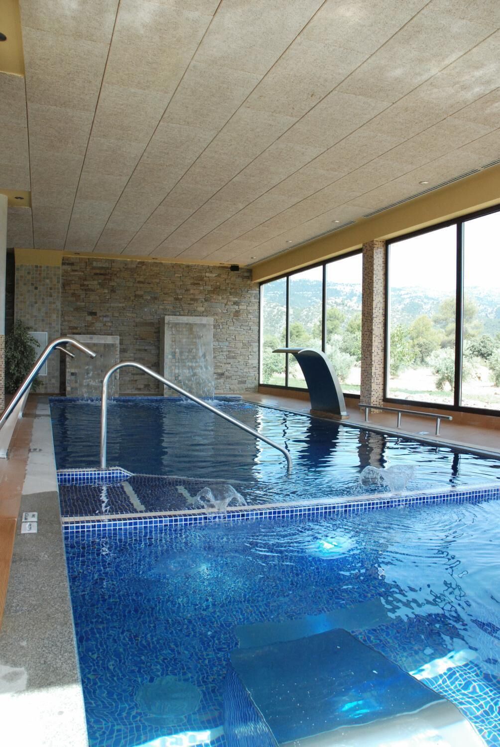An indoor swimming pool with clear water, a stone wall, and a view of the outdoors through large windows.