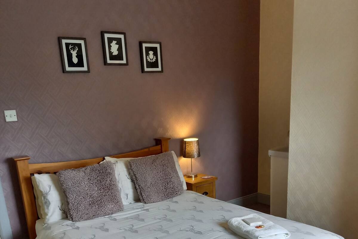 A neatly made bed with a patterned bedspread, two pillows, and two folded towels with a small decorative item on each. There is a bedside lamp on a wooden nightstand beside the bed.