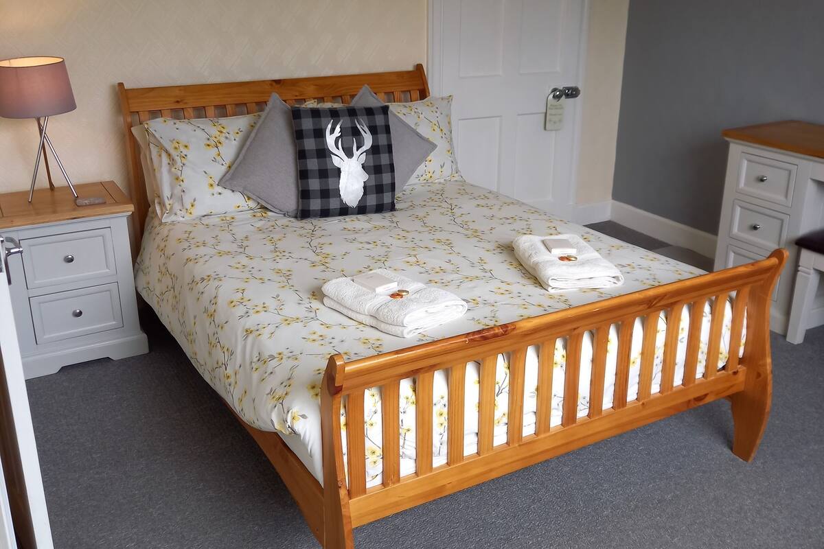 A neatly arranged bedroom with a wooden bed, a bedside table with a lamp, a dresser, and a door with a coat rack.