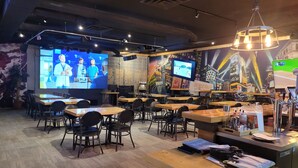 Un restaurante con varias mesas y sillas, una pantalla grande mostrando un noticiero y un mural en la pared.