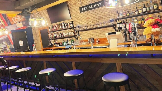 Zona de bar con pared de ladrillo, estantes llenos de botellas de licor y taburetes.