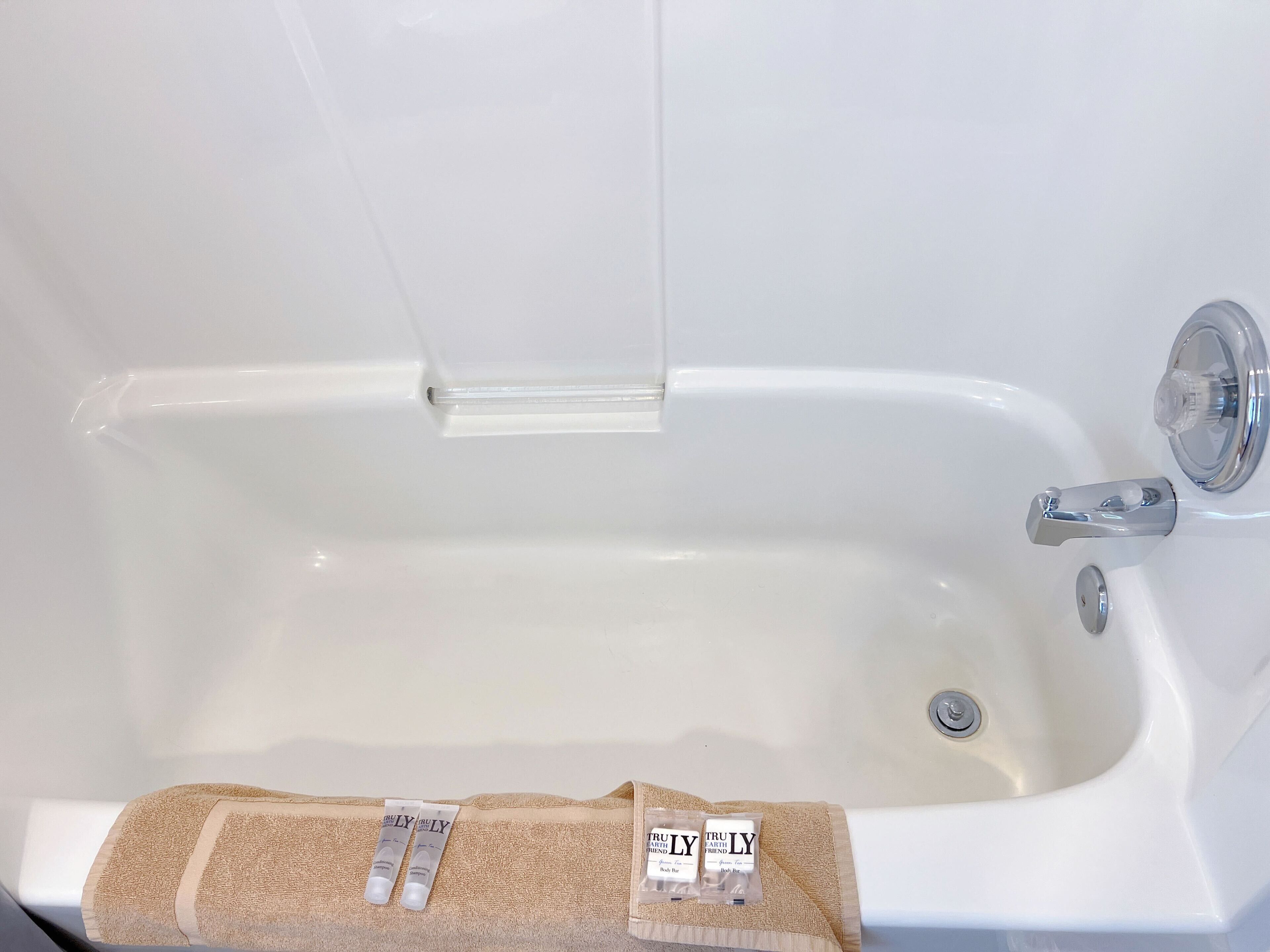 Uma banheira branca com uma torneira de cromo e uma porta de chuveiro de vidro transparente. Há dois tubos de loção sobre uma toalha bege.