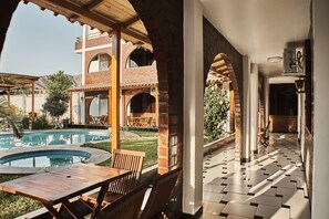 A covered outdoor area with a pool, wooden furniture, and a view of a brick building. - Bris Hotel (Nazca)