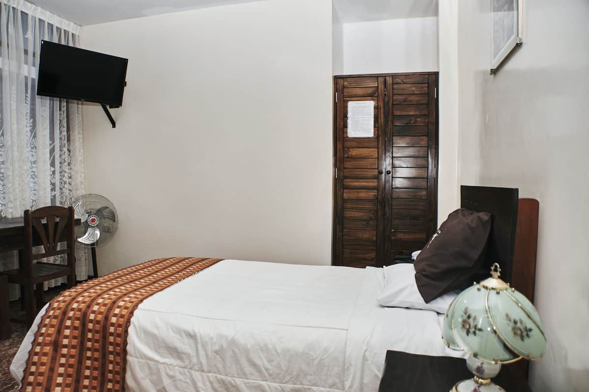 A hotel room with a bed, a desk with a chair, a wall-mounted TV, a wooden wardrobe, and a bedside lamp.