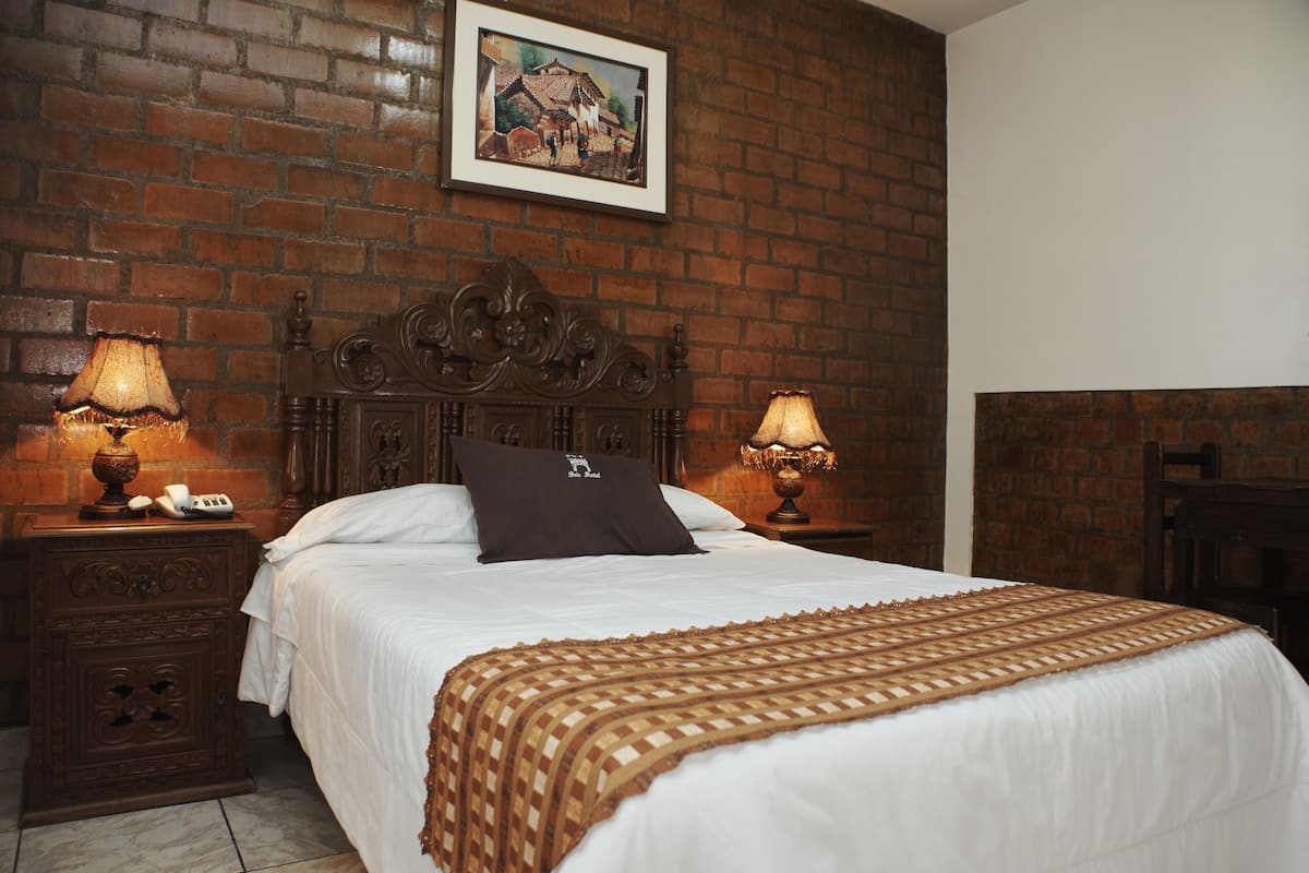A hotel room with a brick wall, a bed with white bedding, two bedside tables with lamps, and a framed picture on the wall.