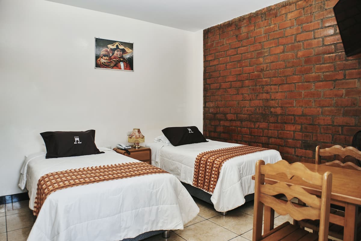 A room with two beds, a brick wall, a wooden table, and a small table with a lamp.