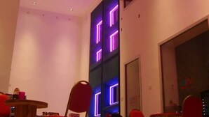 A modern interior with multiple round tables and chairs, a high ceiling with colorful hanging lights, and a large window with purple neon lights. - Hostel Unico (La Plata)