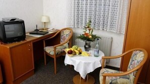 A small hotel room with a desk, a television, two chairs, a table with fruit and a vase, and a window with curtains. - Hotel Restaurant Pempel (Grossalmerode)