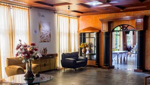 A spacious lobby with a leather sofa, a round glass table, and a vase of flowers. There is a large window with curtains, a wall-mounted artwork, and a wooden archway leading to another room.