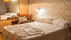 A neatly made bed with white linens and a few towels, a brown armchair with patterned cushions, and a large mirror on the wall.