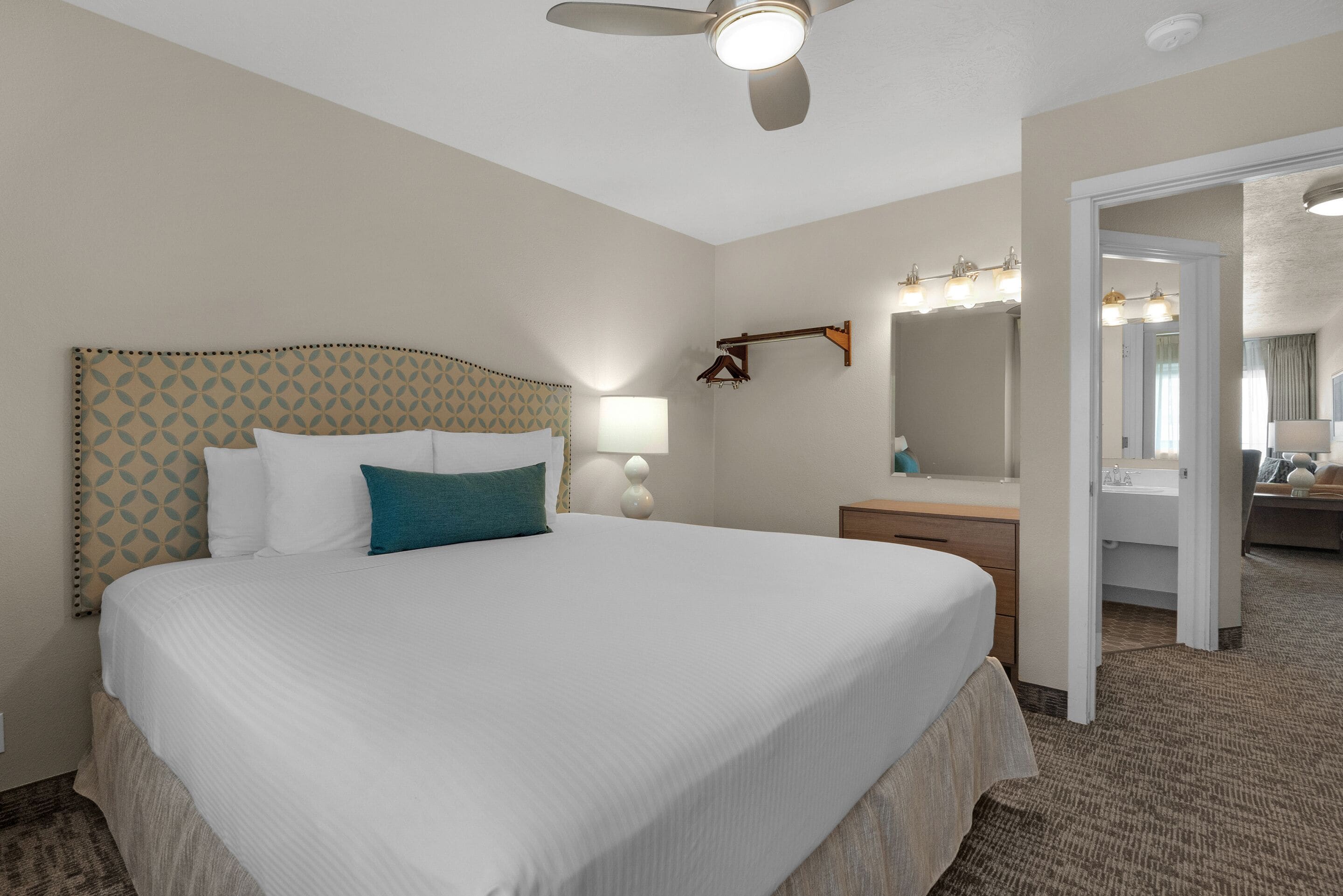 A hotel room with a large bed, a ceiling fan, a vanity with a mirror, and a bathroom visible through an open door.