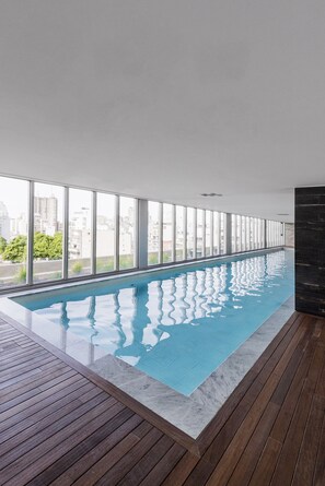 An indoor swimming pool with a wooden deck and large windows offering a city view. - Top Rentals Dorrego (Buenos Aires)