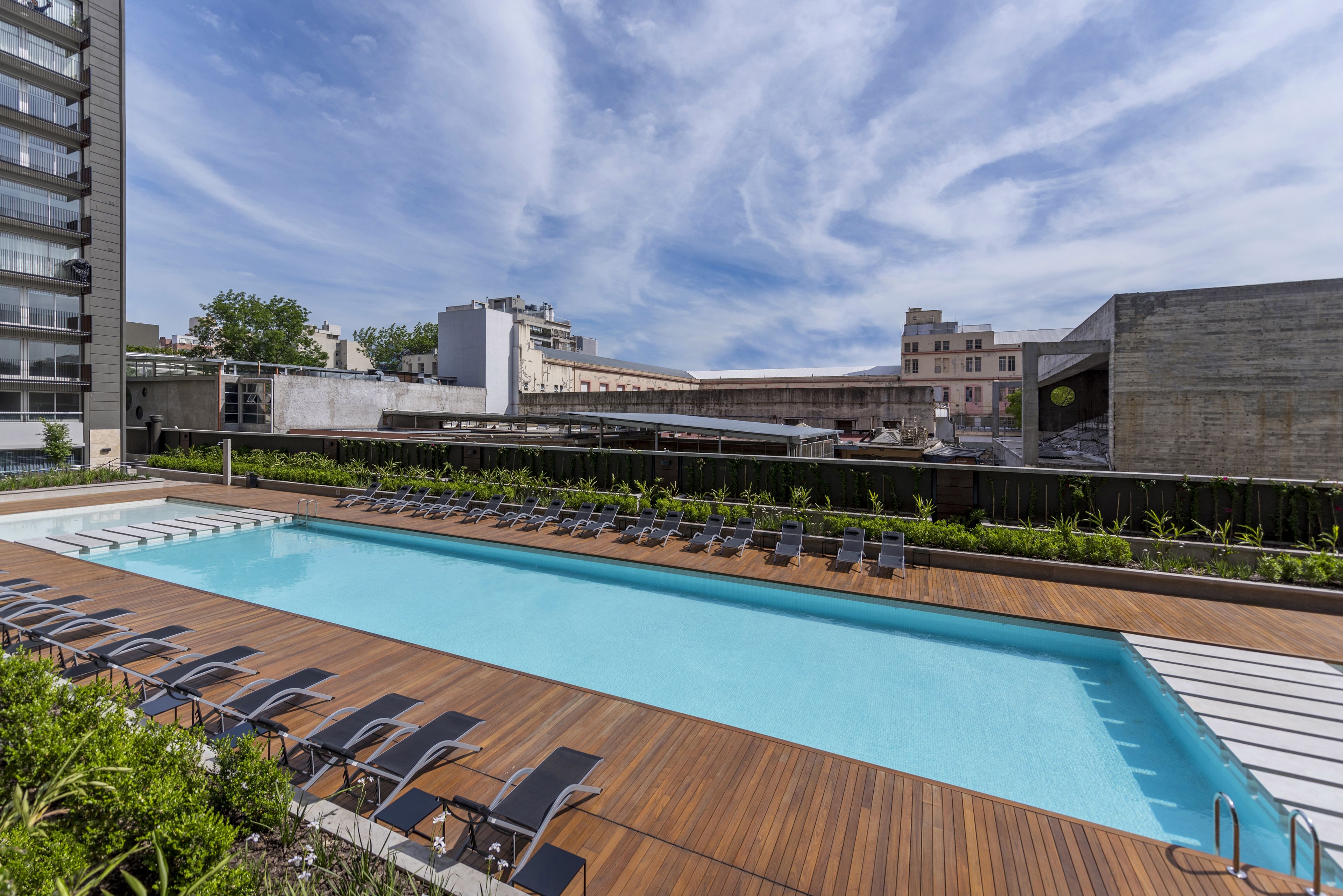 A rooftop swimming pool with wooden deck and lounge chairs, surrounded by buildings.