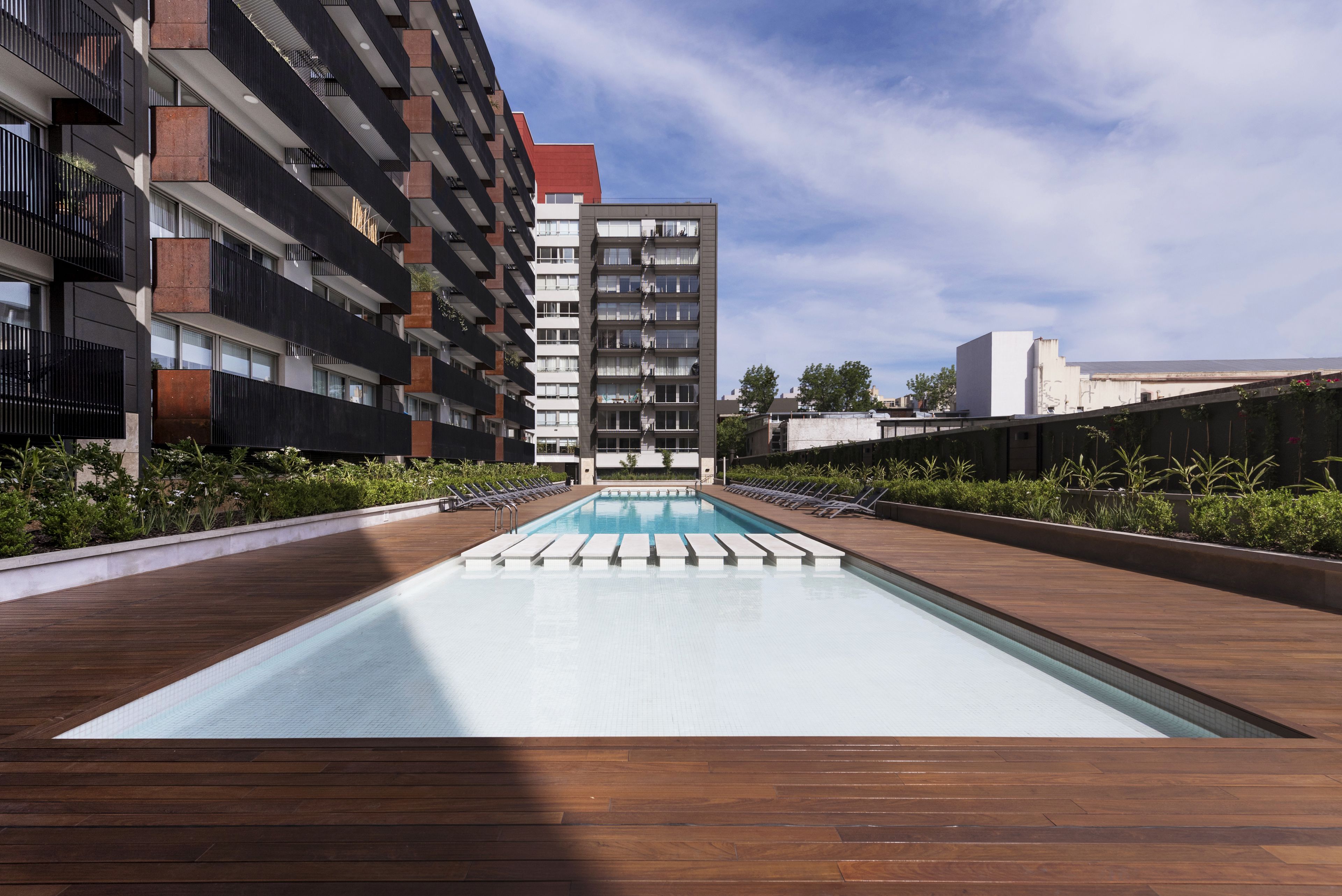 A modern rooftop pool with wooden decking, surrounded by greenery and adjacent to high-rise buildings.