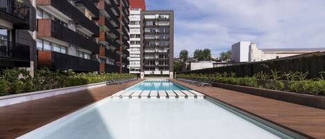 A modern rooftop pool with wooden decking, surrounded by greenery and adjacent to high-rise buildings.