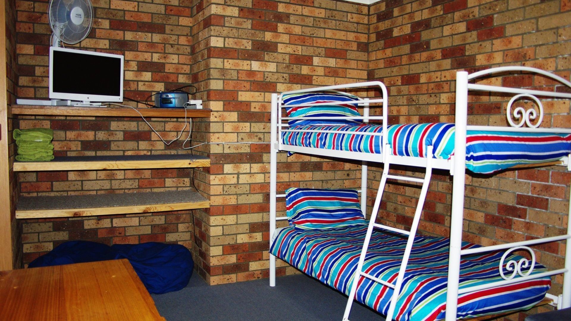A bunk bed with striped bedding, a television, and a fan against a brick wall.