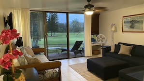 Sala de estar com sofá, poltrona, mesa e ventilador. Janela com vista para um campo de golfe.