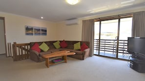 A living room with a brown sofa, colorful cushions, a wooden coffee table, a flat-screen TV, and a balcony with a view. - 2/21 Montague Street - Boat Parking (BERMAGUI)