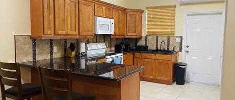 Cocina compacta con armarios de madera, microondas, estufa y pileta. Hay dos taburetes de bar y un bote de basura.