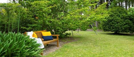 A wooden bench with cushions, surrounded by lush greenery and trees.