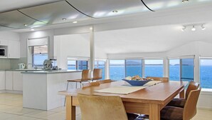 Uma cozinha moderna com uma ilha grande, uma área de refeições com vista para o mar, e uma sala de estar espaçosa com janelas amplas.