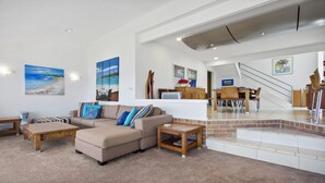 Uma sala de estar ampla com sofá, mesa de centro e mesa de jantar com cadeiras. Há uma escada que leva a um andar superior.