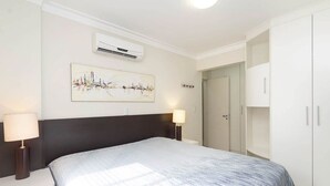 A bedroom with a bed, bedside lamps, a wall-mounted air conditioner, a door, and a wardrobe. - Vila Maior 207 - Praia de Bombas (Bombinhas)