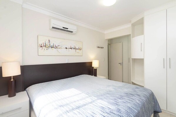 Quarto com cama, luminárias de cabeceira, ar condicionado na parede, porta e guarda-roupa.