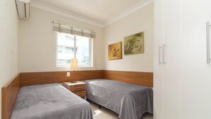A room with two beds, a window with blinds, a lamp, and two wall-mounted paintings. - Vila Maior 207 - Praia de Bombas (Bombinhas)