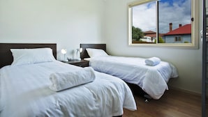 A hotel room with two beds, white linens, bedside lamps, a window with a view of houses, and a wooden floor.