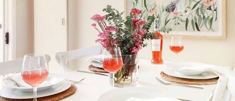 Ein Esstisch ist gedeckt mit weißen Tellern, klaren Weingläsern und einer Vase mit rosafarbenen Blumen.